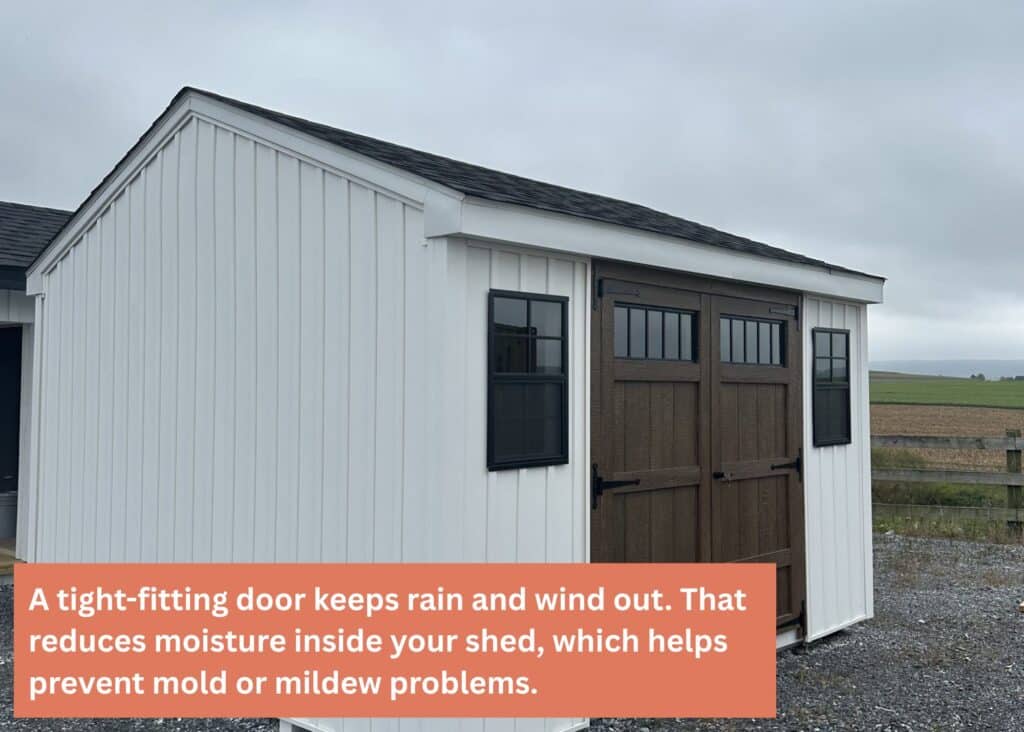 White board-and-batten shed with a dark brown double door and black-trimmed windows, sitting on a gravel lot under an overcast sky. Caption: A tight-fitting door keeps rain and wind out. That reduces moisture inside your shed, which helps prevent mold or mildew problems.