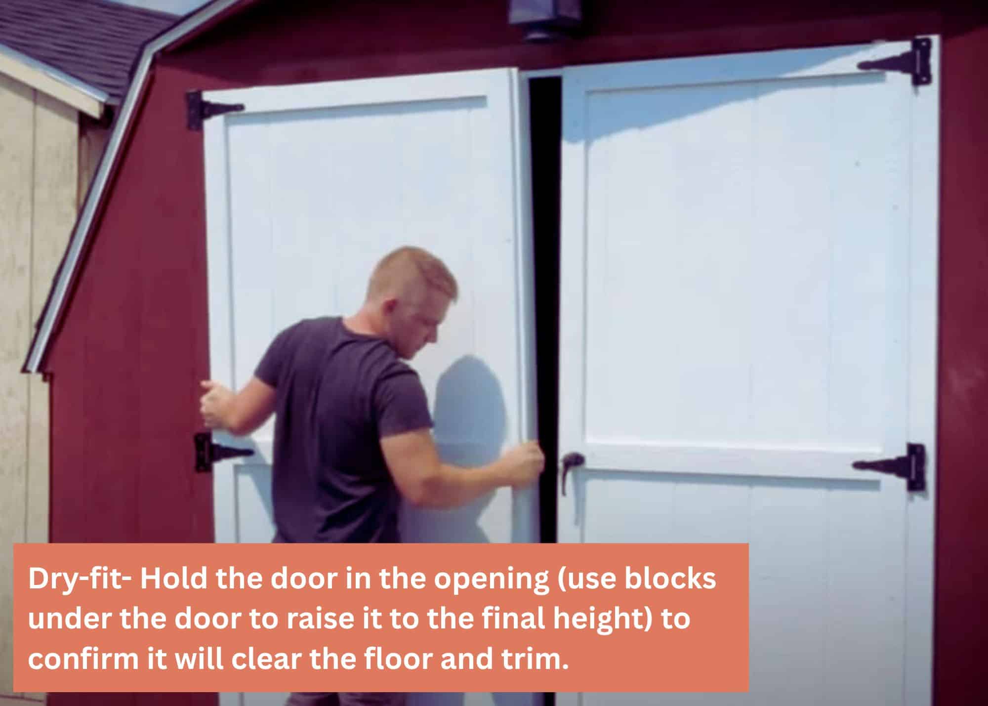 Man installing or adjusting white double doors on a red wooden shed, with another tan shed visible in the background under a clear sky. Caption: Dry-fit- Hold the door in the opening (use blocks under the door to raise it to the final height) to confirm it will clear the floor and trim.