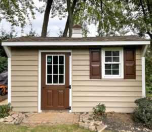 Tan vinyl shed with a brown single door featuring window panes, matching brown shutters, and a small cupola on the roof, surrounded by landscaping. fiberglass-shed-doors-with-glass-for-shed-in-pennsylvania